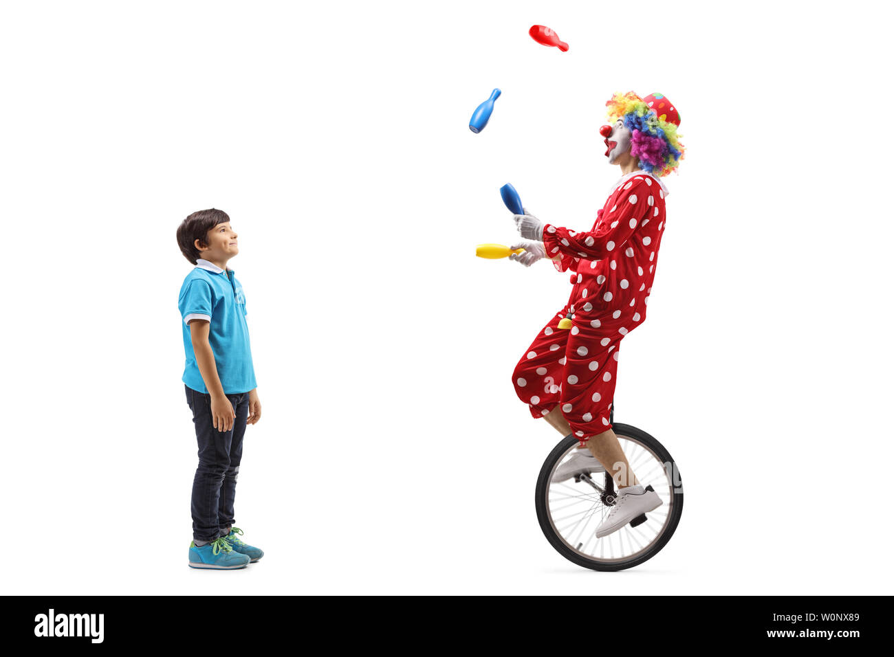 Full length profile shot of a boy watching a clown juggling and riding a unicycle isolated on white background Stock Photo