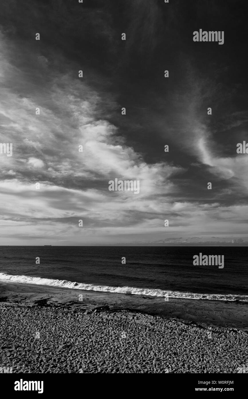 Black and white image of a beach with a solitary coal tanker on the horizon beneath a moody cloudy sky Stock Photo