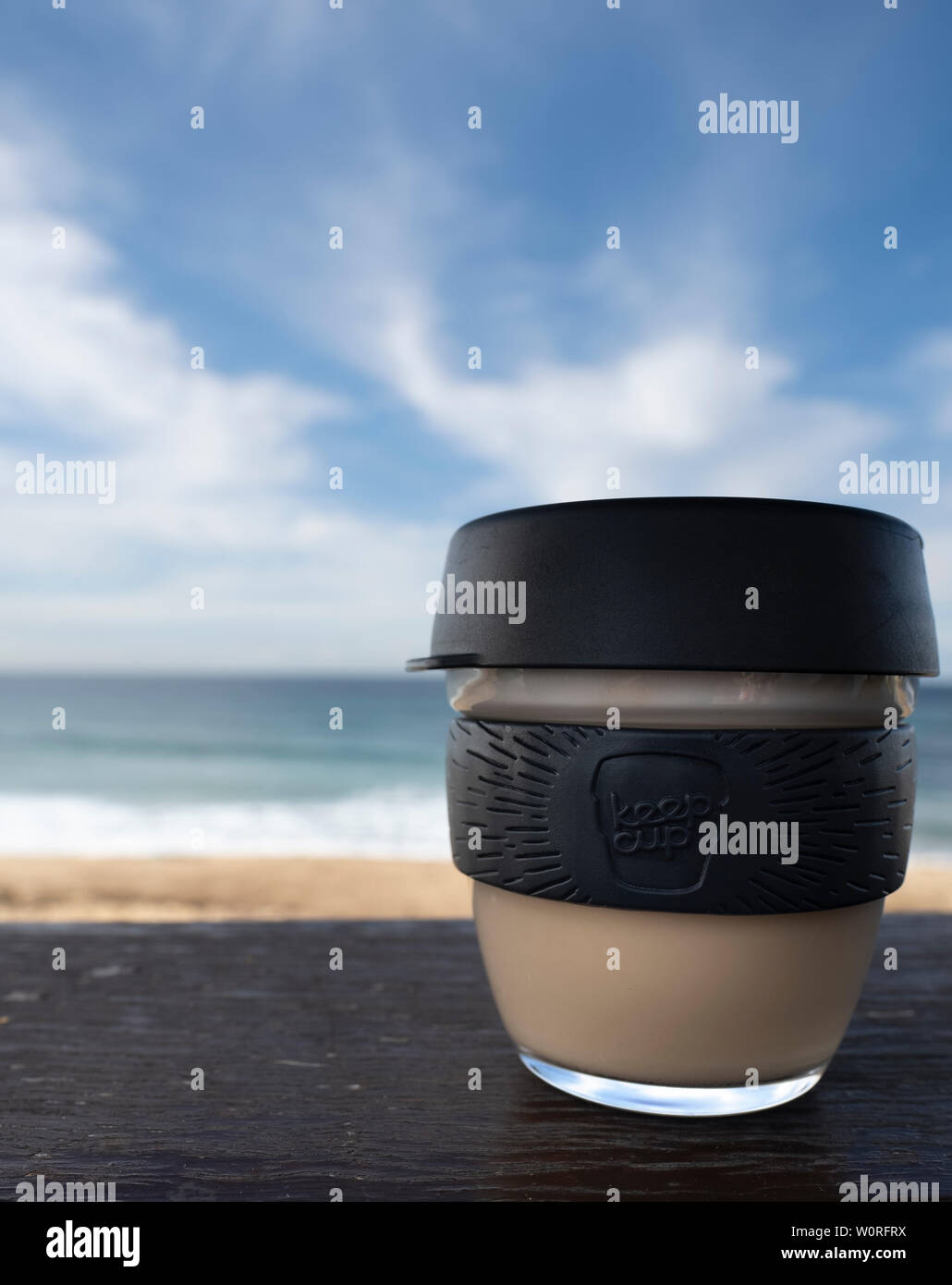 NEWCASTLE, AUSTRALIA - June 16 2019 Close up of a glass Keepcup with black trimming, filled with coffee, on a timber bench with the Pacific Ocean in t Stock Photo