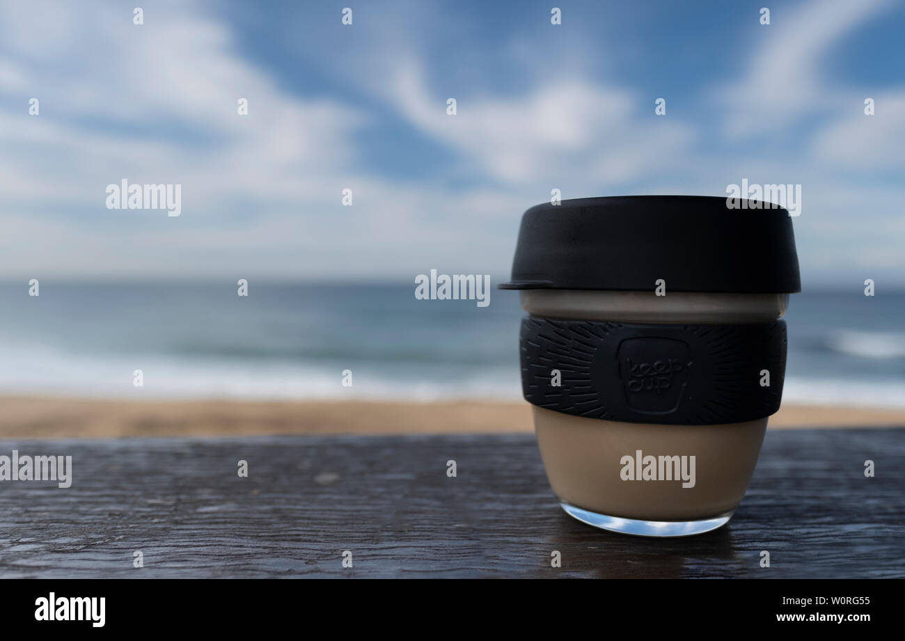 NEWCASTLE, AUSTRALIA - June 16 2019 Close up of a glass Keepcup with black trimming, filled with coffee, on a timber bench with the Pacific Ocean in t Stock Photo