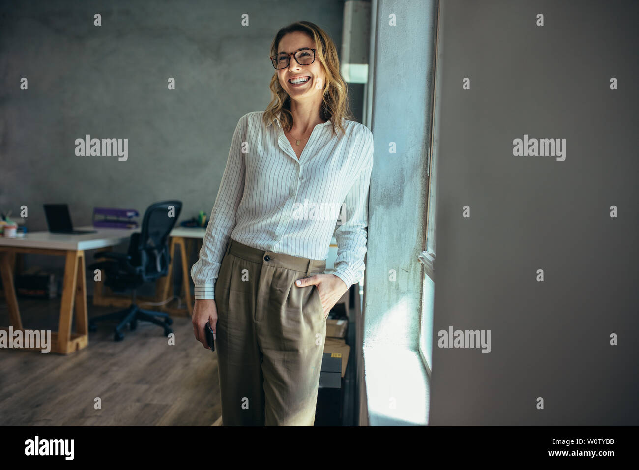 Smiling businesswoman standing in office. Online business owner in casuals standing by window in office looking away and smiling. Stock Photo
