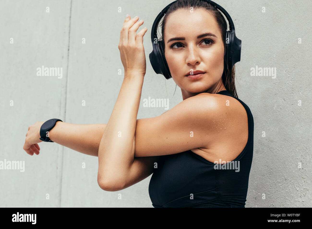 Fit young woman stretching her arms outdoors. Woman in sportswear and heaphones doing warmup exercise in the morning against grey wall. Stock Photo