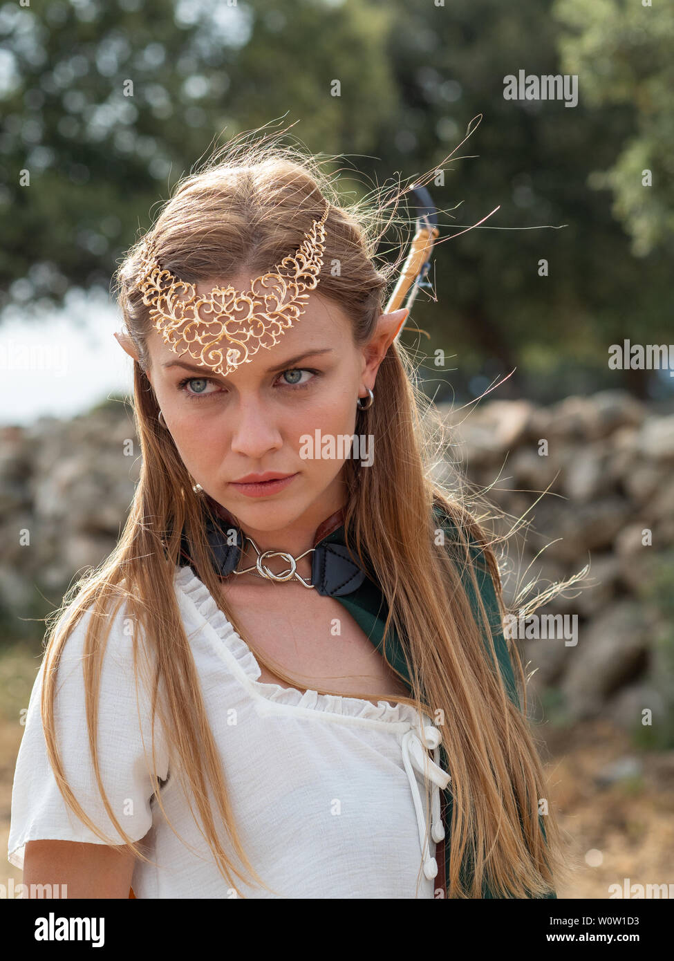 Blonde girl with blue eyes and makeup with elf ears poses in the field with  a bow and a green cape Stock Photo - Alamy