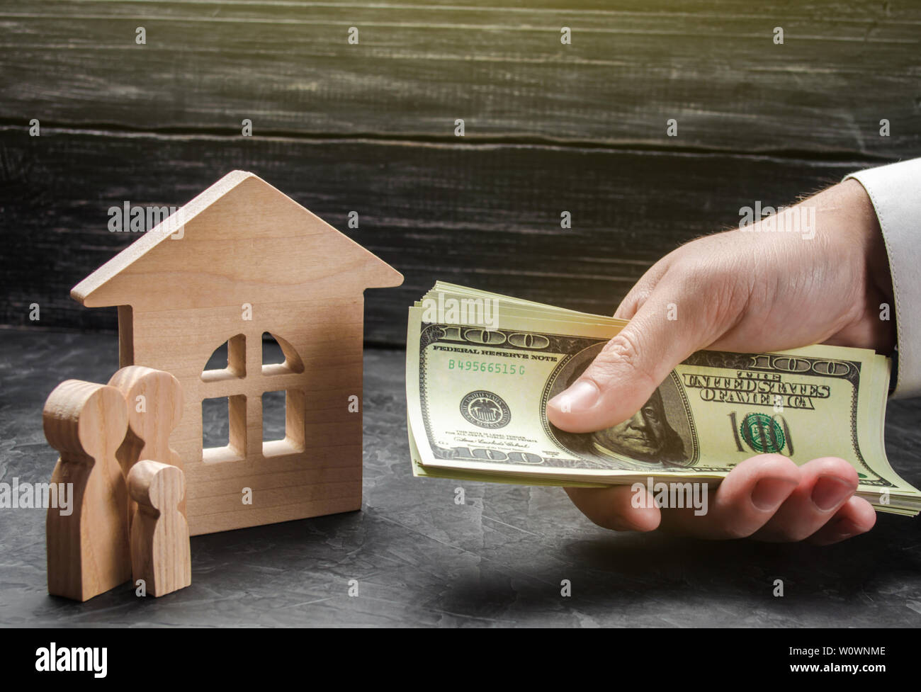 The hand of a businessman stretches a wad of money to the family figures near the wooden house. Providing a mortgage loan for buying a home and improv Stock Photo