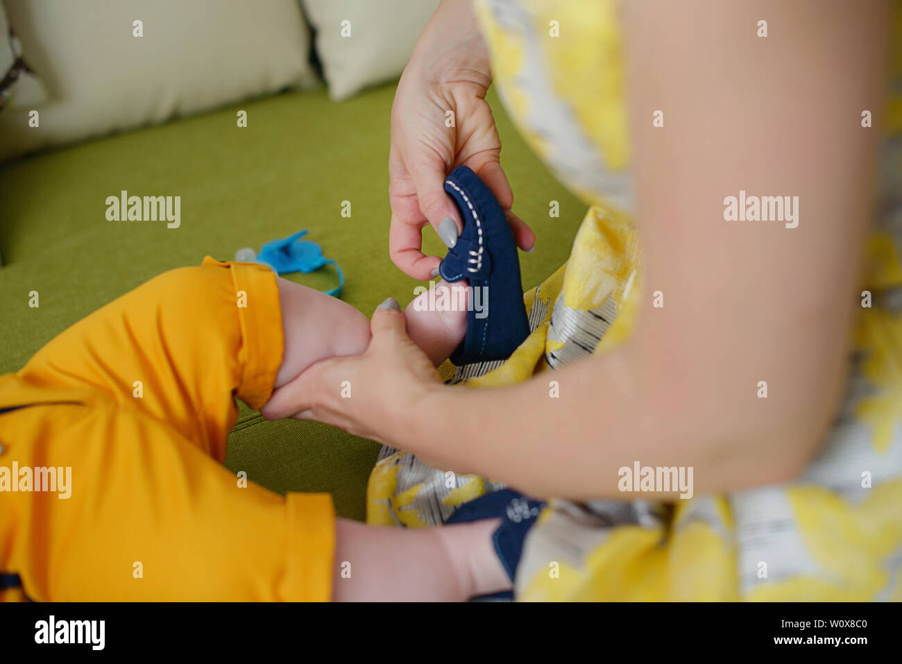 Caucasian young mother wearing a vintage yellow dress and helping little baby boy getting ready, with focus on the hands fitting cute blue shoes Stock Photo
