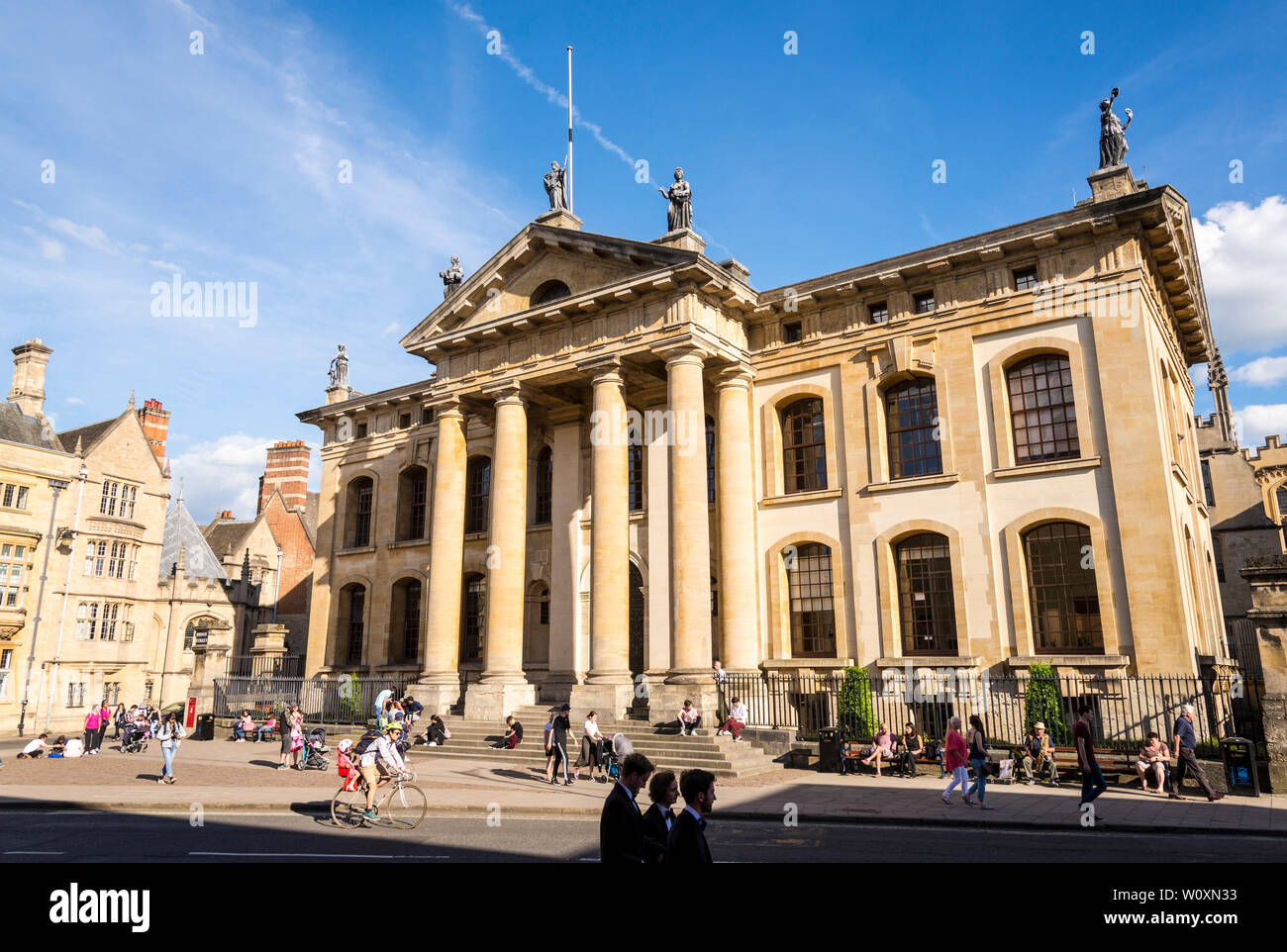 People sit on the steps at the front of the Clarendon Building brightly sunlit on a beautiful summery day in the famous university town of Oxford Stock Photo