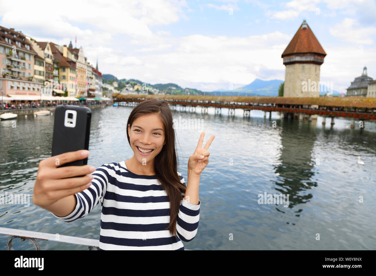 Tourist selfie woman taking self portrait photograph picture with smartphone in Lucerne Switzerland. Travel woman with smart phone by Kapellbrucke Chapel Bridge and Wasserturm water tower, Reuss River Stock Photo
