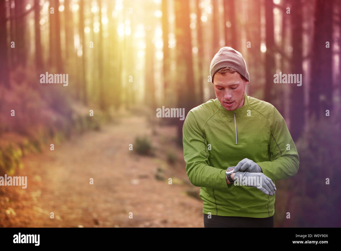 Trail running runner looking at heart rate monitor watch running in forest wearing warm jacket sportswear, hat and gloves. Male jogger running training in woods. Stock Photo