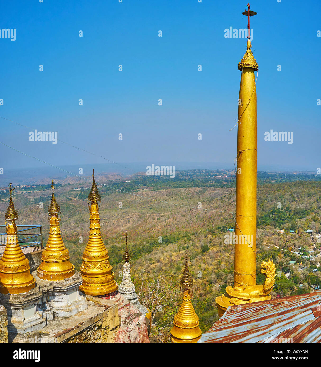 The small golden stupas and tall slender Buddhist pillar in Taung Kalat monastery  on the edge of Popa mountain outcrop, with a view on green hills og Stock Photo