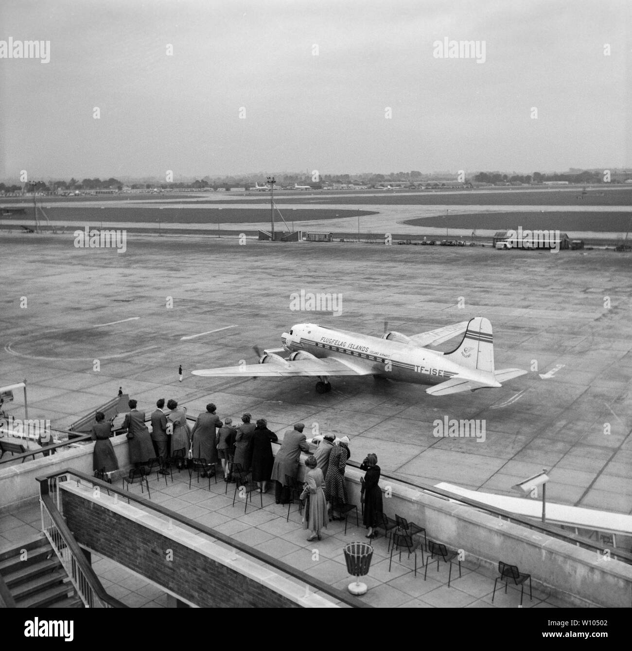 A Vintage 1950s black and white photograph taken at London Heathrow Airport, showing a group of people standing and looking over a balcony towards a Flugfelag (Iceland Airways), Douglas C-54 (DC-4) airliner. Stock Photo