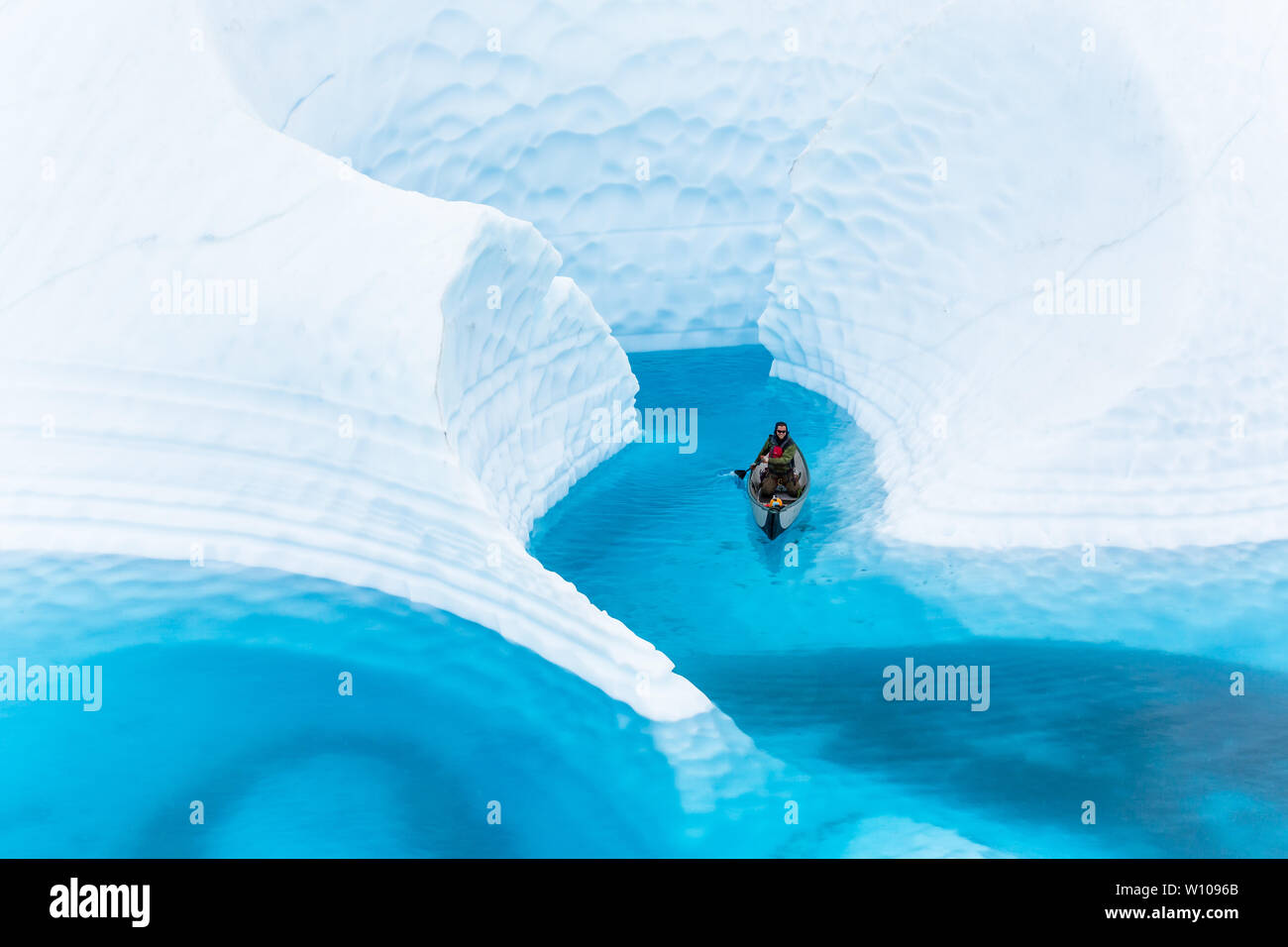 Tall walls of white glacier ice surround a young man in a boat, paddling the deep blue water of a supraglacial lake in remote Alaska. Stock Photo