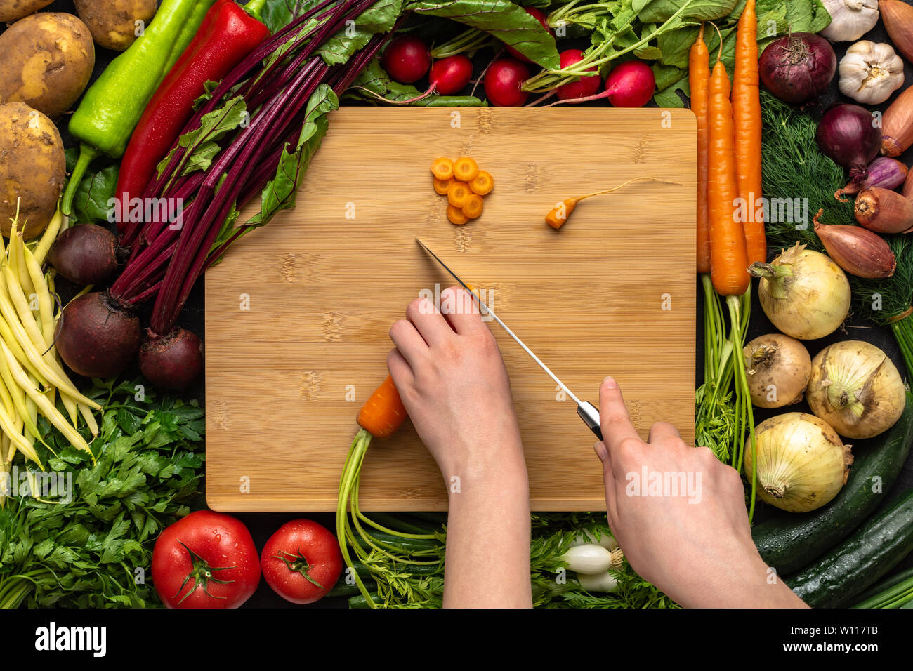 Female Hands Cutting Organic Carrot with Chef Knife on Wooden Chopping Board. Healthy Vegetarian Food Concept. Stock Photo