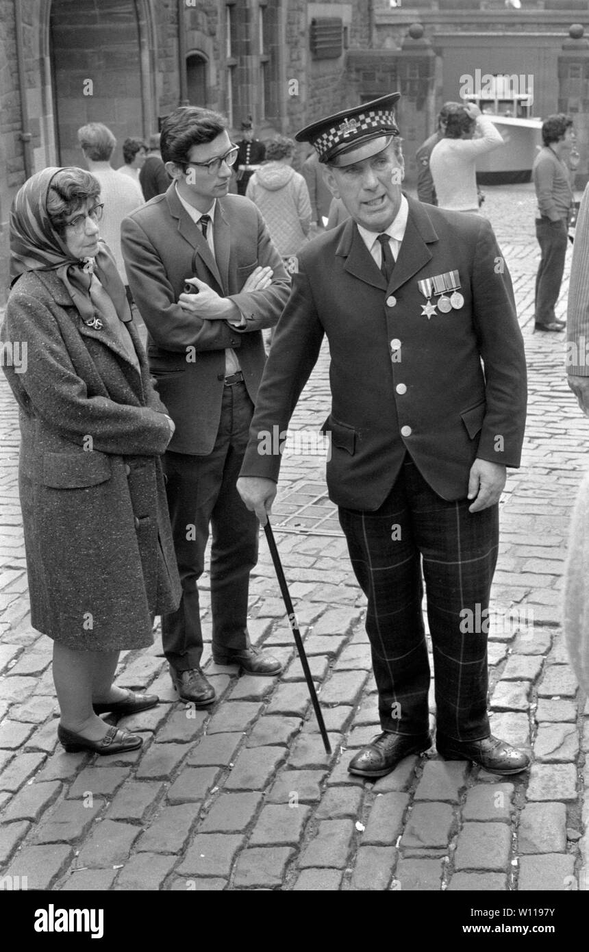 Elderly old soldier wearing medals works as tour guide group people Edinburgh Castle 1970s Scotland UK   1971 HOMER SYKES Stock Photo