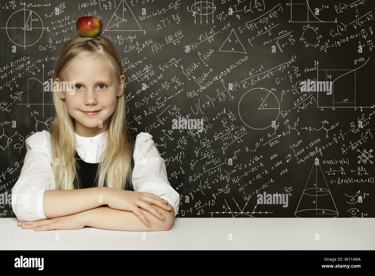 Cute child student girl with red apple on head. Blackboard background with science formulas. Learning science concept. Stock Photo