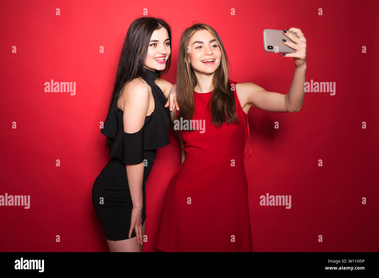 Two women make take selfies on phone wear dresses isolated red background Stock Photo