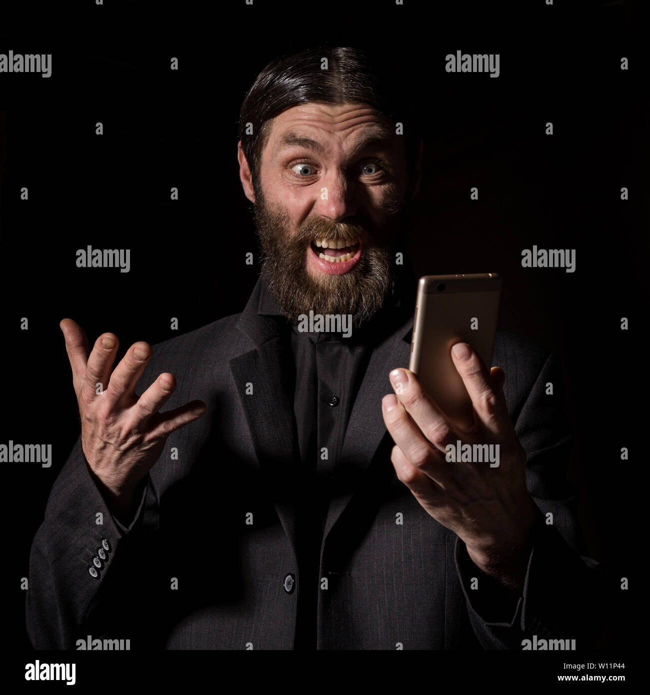 Old believer senior priest with a smartphone, bearded old man is calling on a dark background. Stock Photo
