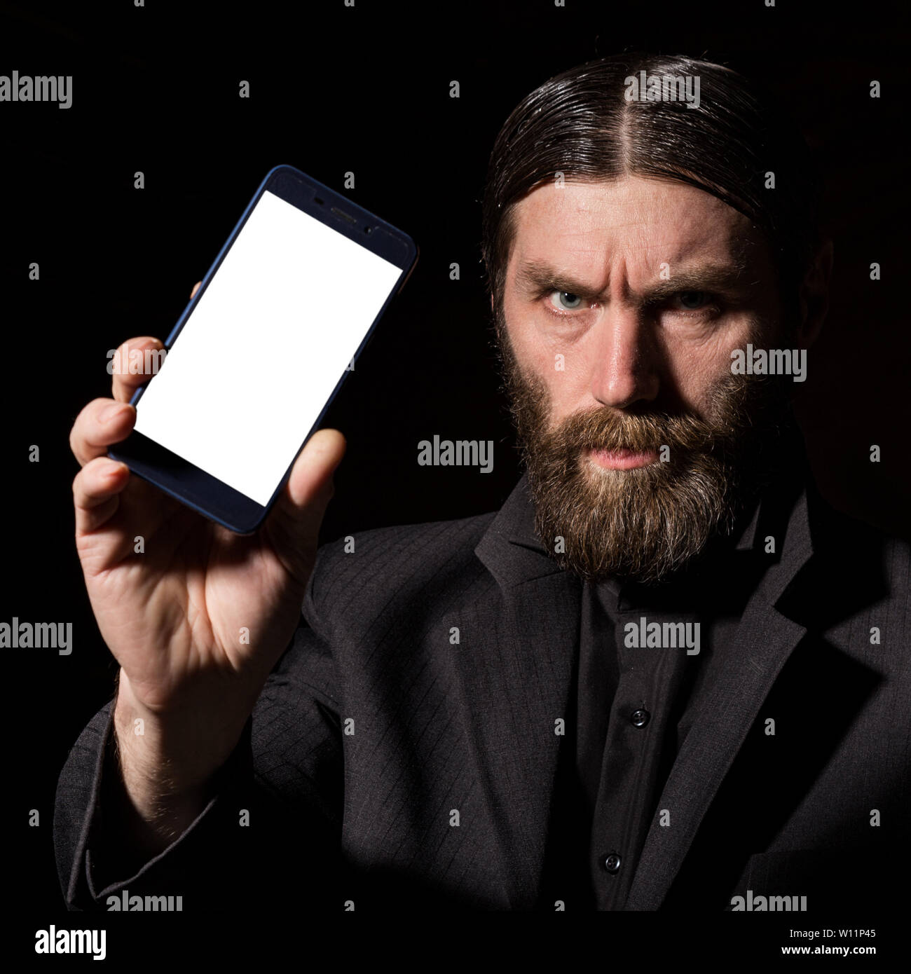 Old believer senior priest with a smartphone, bearded old man is calling on a dark background. Stock Photo