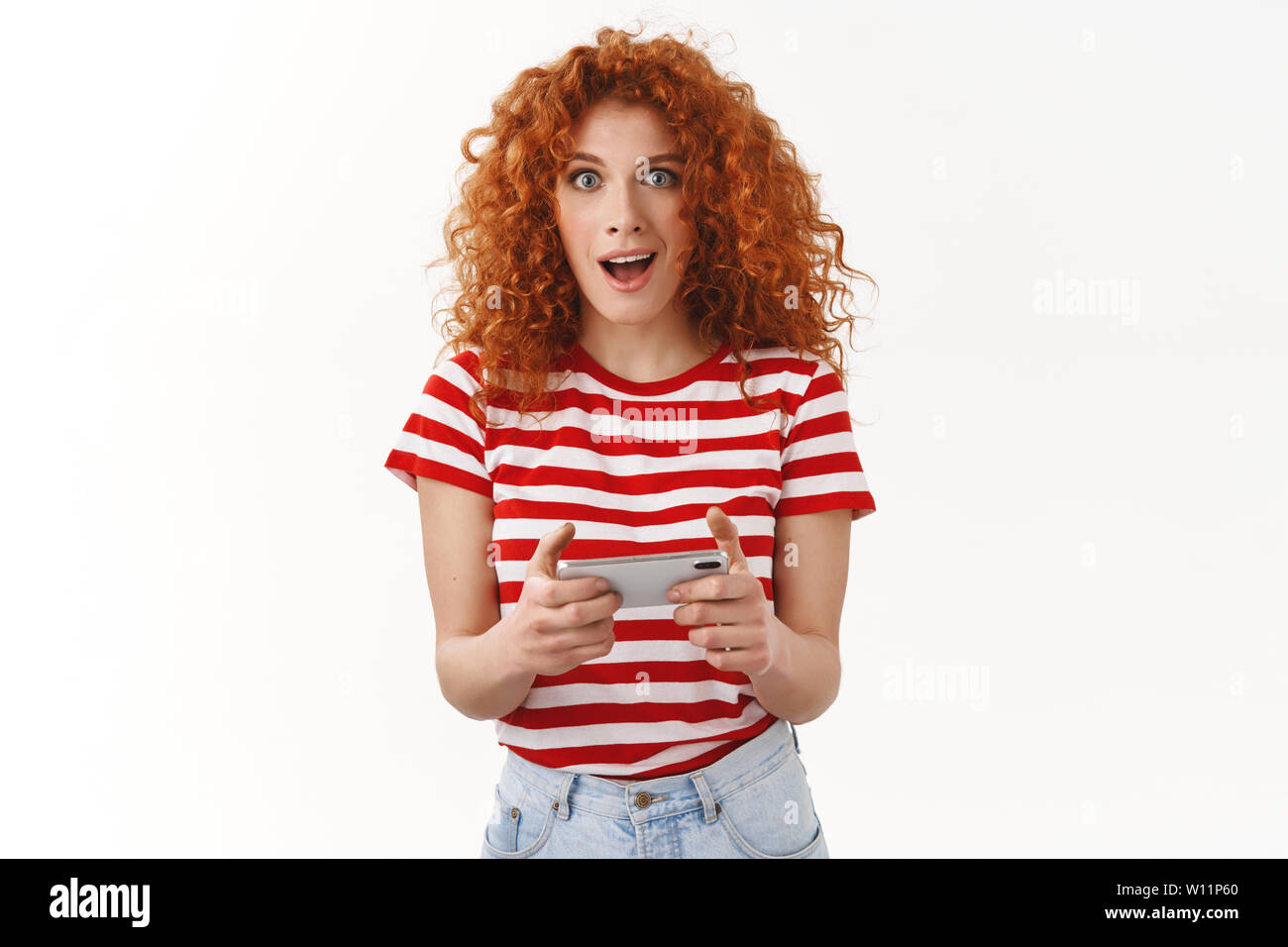 Amused excited good-looking female redhead geek attractive woman playing cool new app smartphone game look camera surprised winning first place standi Stock Photo