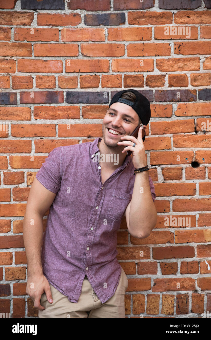 Handsome Young Caucasian Man with Backwards Hat Smiling for Portraits in Front of Textured Brick Wall Outside Stock Photo