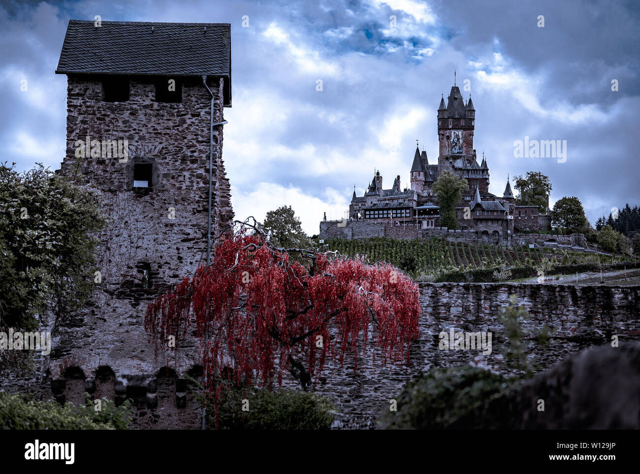 Dramatic fairytale castle on a hill with dark sky clouds and red tree. Stock Photo