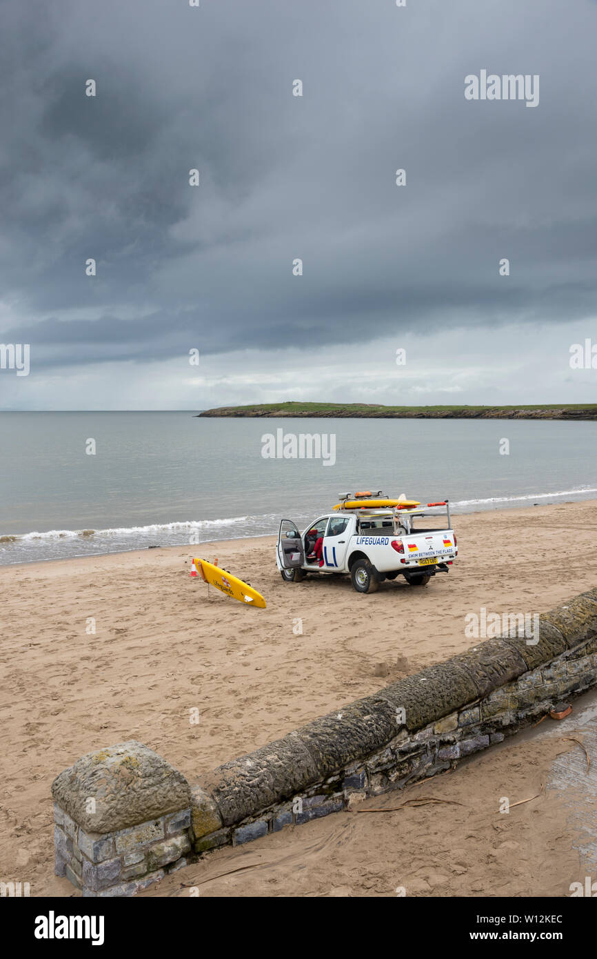 A lifeguard van is parked on the sandy beach near the shoreline. In the background a headland juts into the calm sea  beneath dark rain clouds. Stock Photo