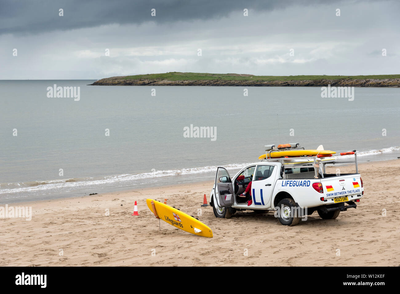 A lifeguard van is parked on the sandy beach near the shoreline. In the background a headland juts into the calm sea  beneath dark rain clouds. Stock Photo