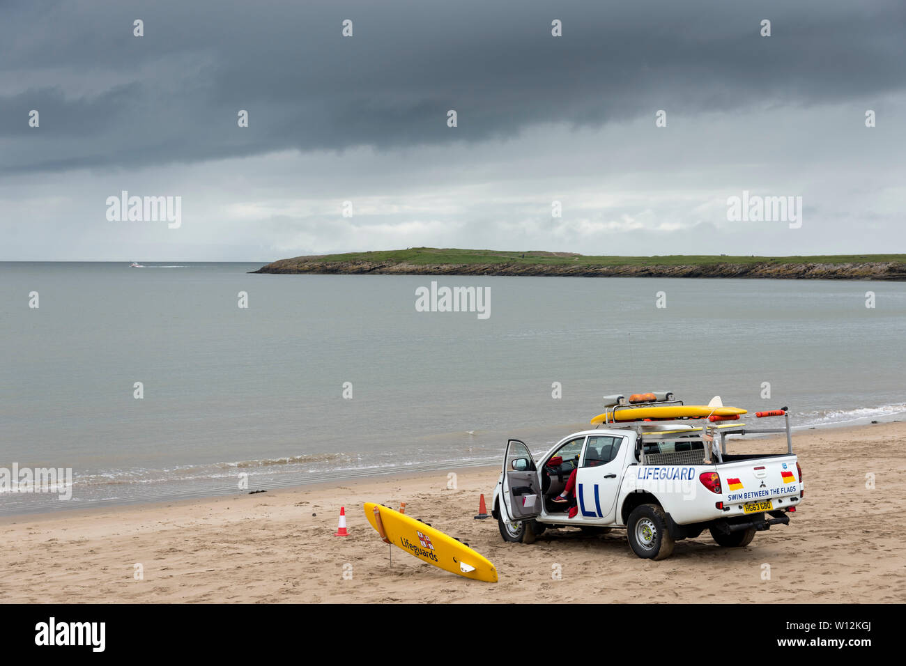 A lifeguard van is parked on the sandy beach near the shoreline. In the background a headland juts into the calm sea  beneath dark rain clouds. Stock Photo