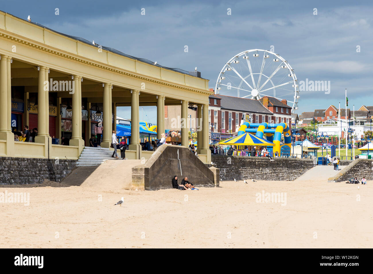 The sandy beach and Ferris wheel at Barry Island are brightly lit by sunshine and stand out against the dark clouds in the sky on an early summer day Stock Photo