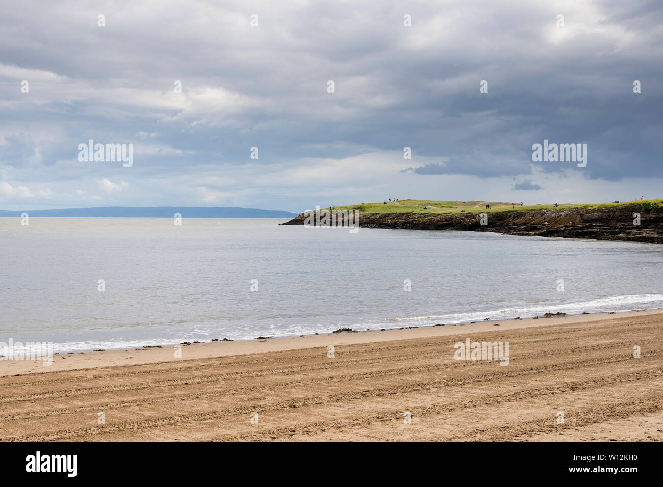 Sun shines during a break in the clouds onto Barry Island's sandy beach, striped by tractor tyre tracks, and the low-lying Friar's point headland. Stock Photo