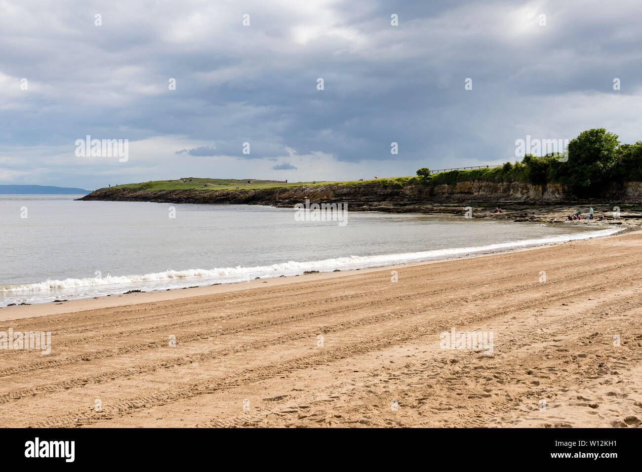 Sun shines during a break in the clouds onto Barry Island's sandy beach, striped by tractor tyre tracks, and the low-lying Friar's point headland. Stock Photo