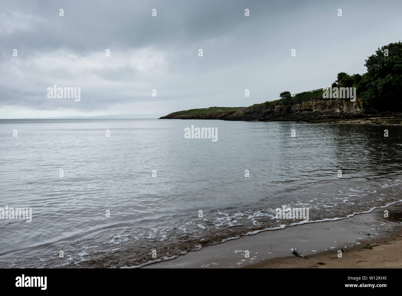 The sea is calm an smooth and barely causes a ripple on the shoreline. In the background Friar's Point is a dark headland under a stormy sky. Stock Photo