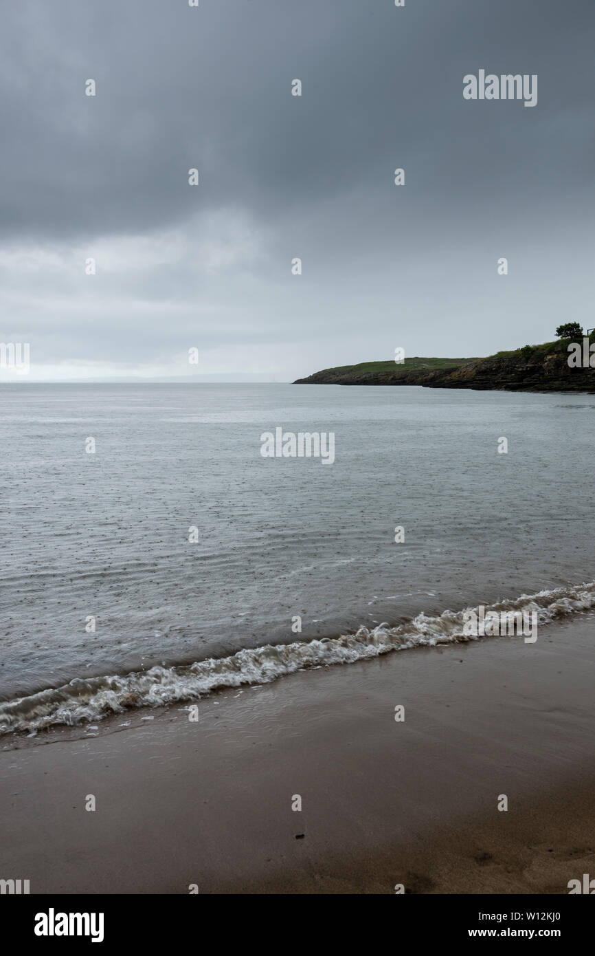 The sea, pocked marked by rain during a heavy shower, is calm and laps gently on the beach. In the background is a dark headland under a stormy sky. Stock Photo