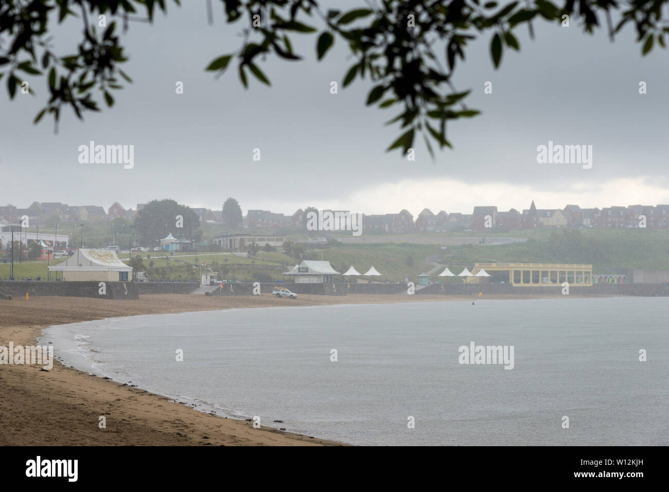 Tree foliage frames a view of the sandy beach at Barry Island which is empty, but for a distant lifeguard van, during a heavy shower. Stock Photo