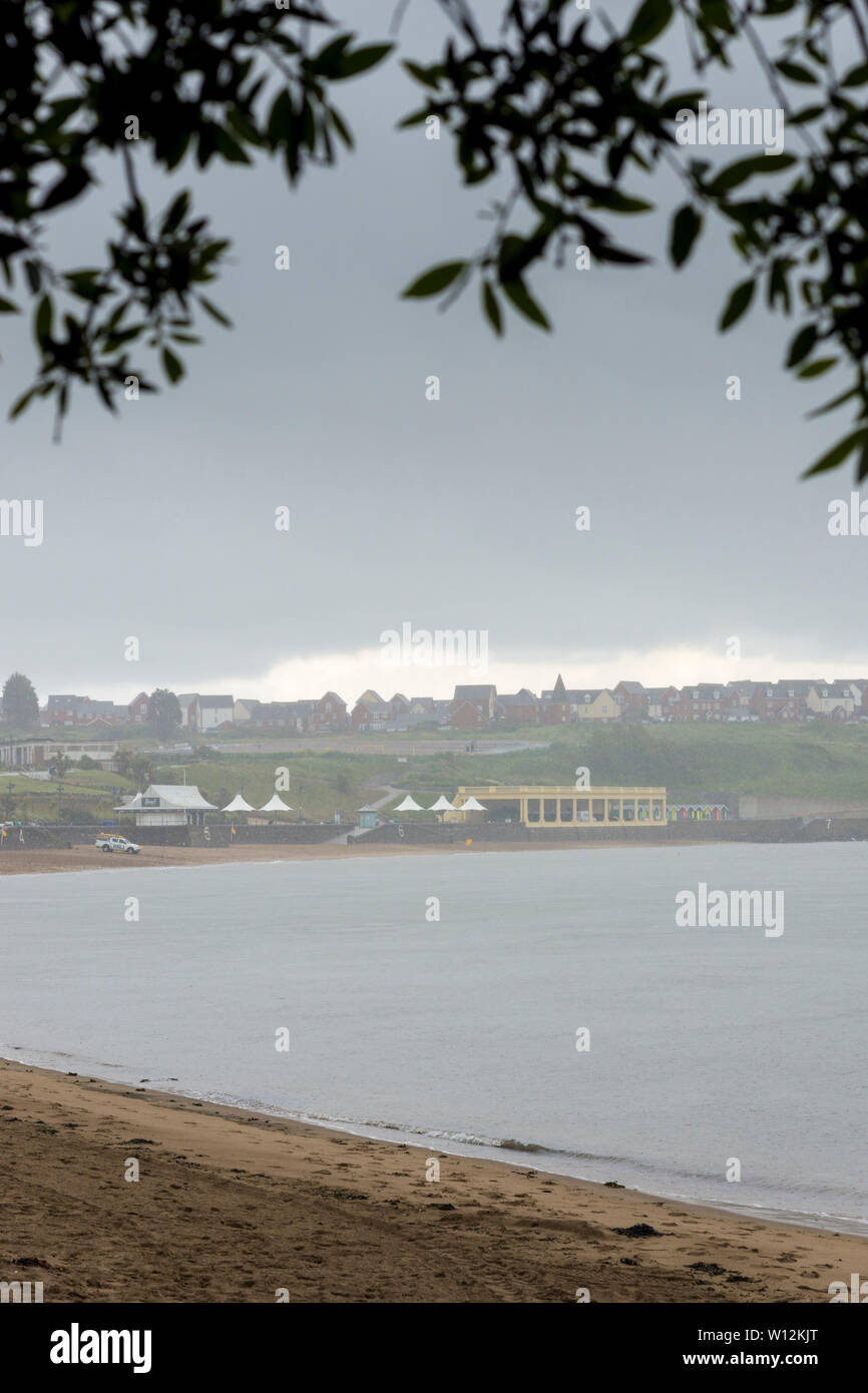 Tree foliage frames a view of the sandy beach at Barry Island which is empty, but for a distant lifeguard van, during a heavy shower. Stock Photo