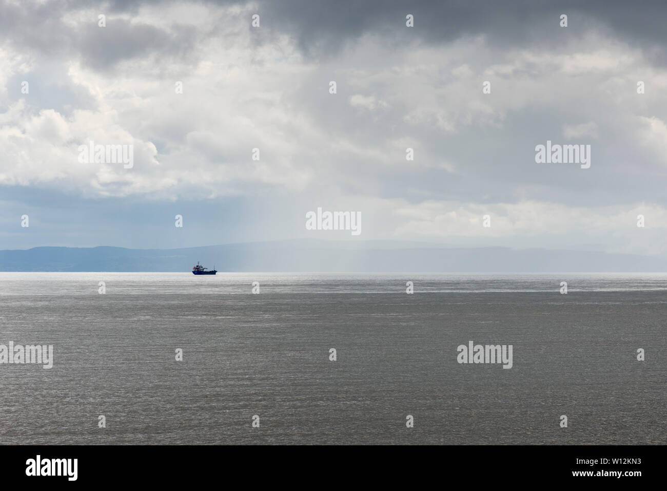 In the distance a ship sits in a patch of sunshine on the Bristol Channel on a cloudy day with coast of Somerset behind it. Stock Photo