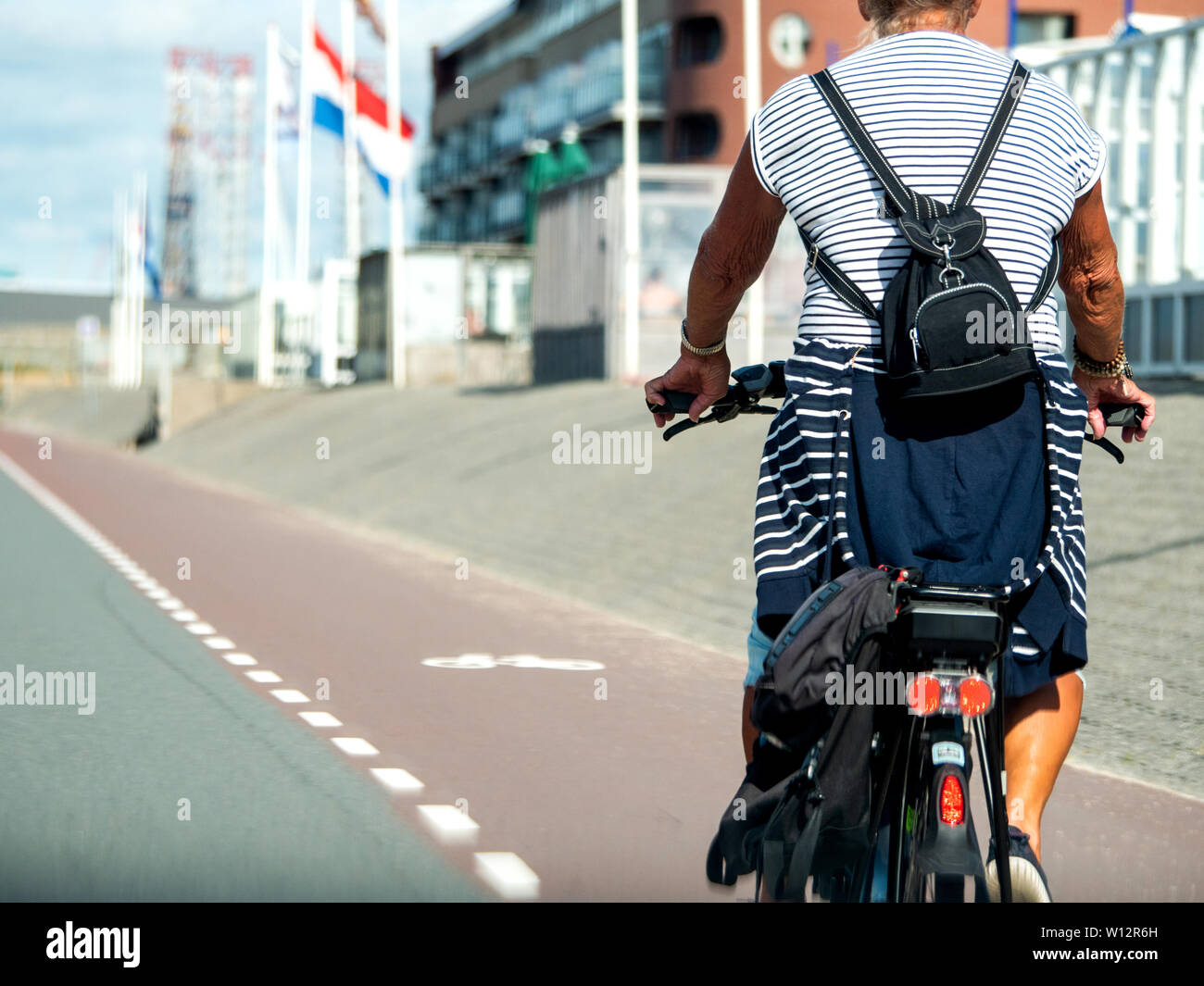 Rear view of unrecognizable adult woman riding a bicycle on the dedicated cycling lane in Netherland with Dutch flag in the background Stock Photo