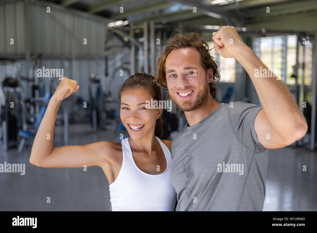 Fit power couple happy flexing strong arms showing off success training at fitness gym - Smiling Asian woman, Caucasian man . Stock Photo