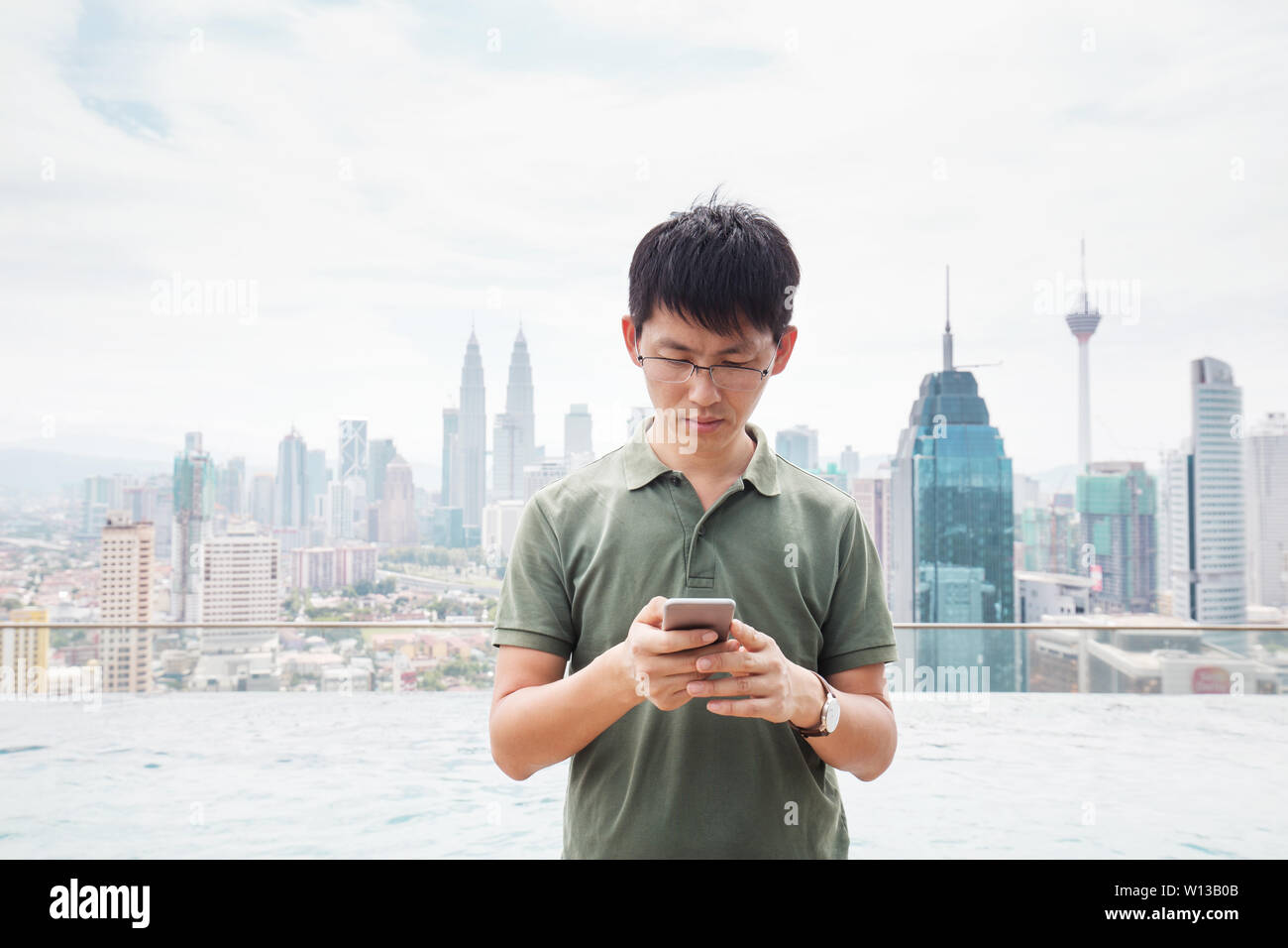 young chinese man with mobile phone stands by swimming pool on balcony in midtown of kuala lumpur in cloud sky Stock Photo