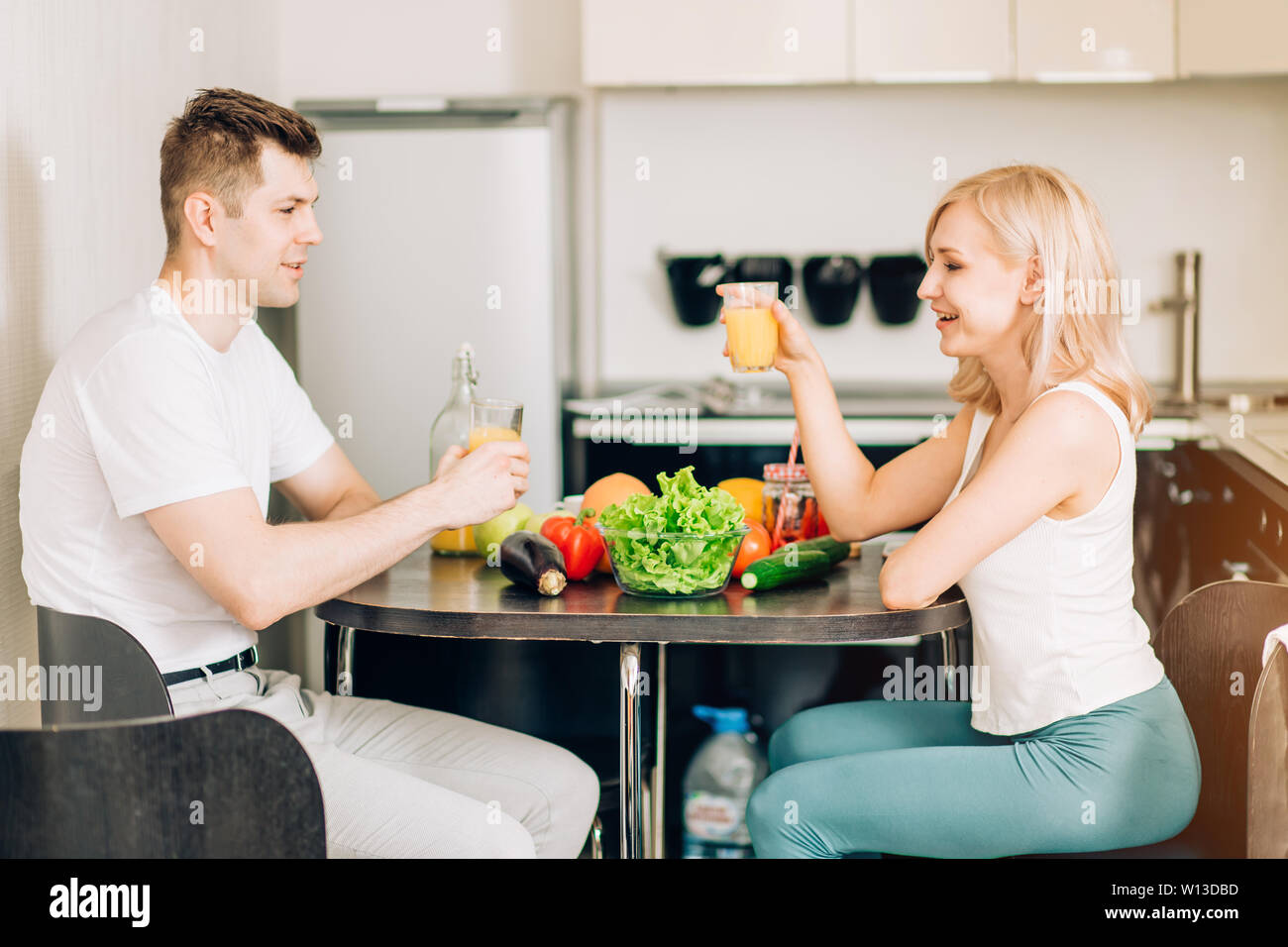 Beautiful young couple is talking, looking at each other and smiling while having breakfast, drinking fresh orange juice, having a great time together Stock Photo