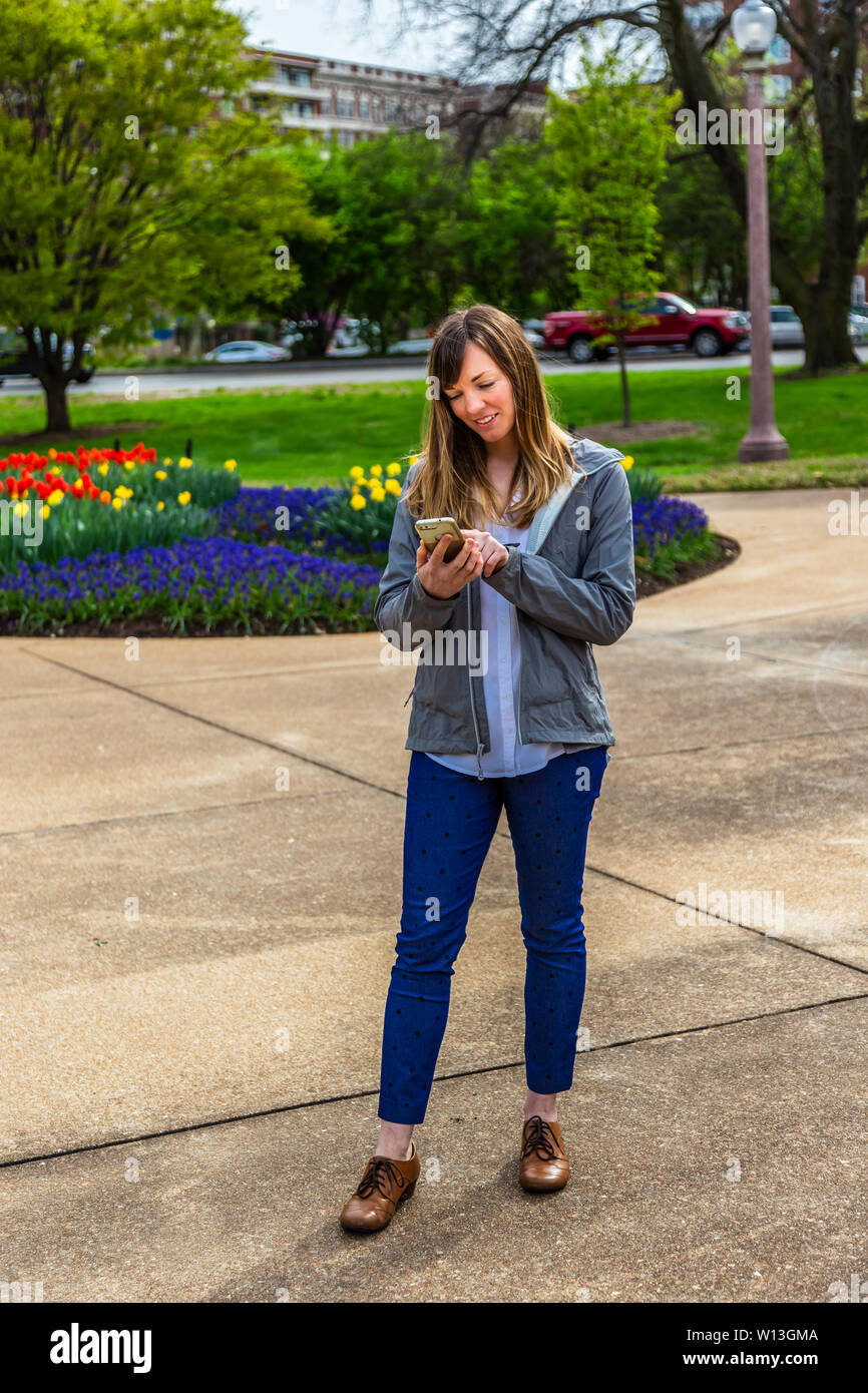 Young woman standing looking down flowersat her phone in a park. Stock Photo