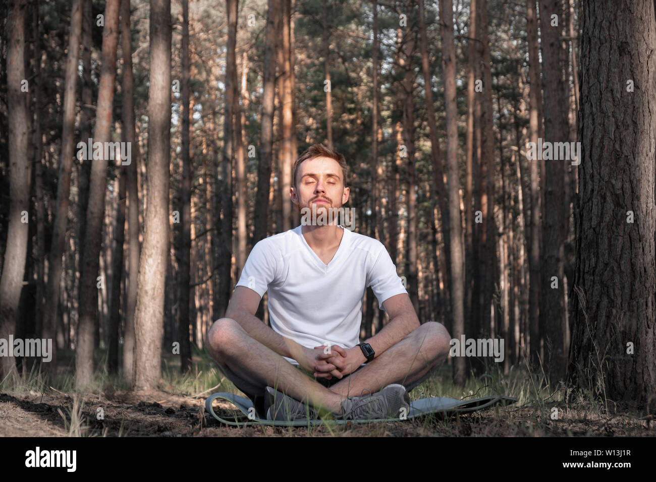 Young male person meditating in the forest. Modern man sits in the pine woods with closed eyes and enjoys the silence of nature Stock Photo
