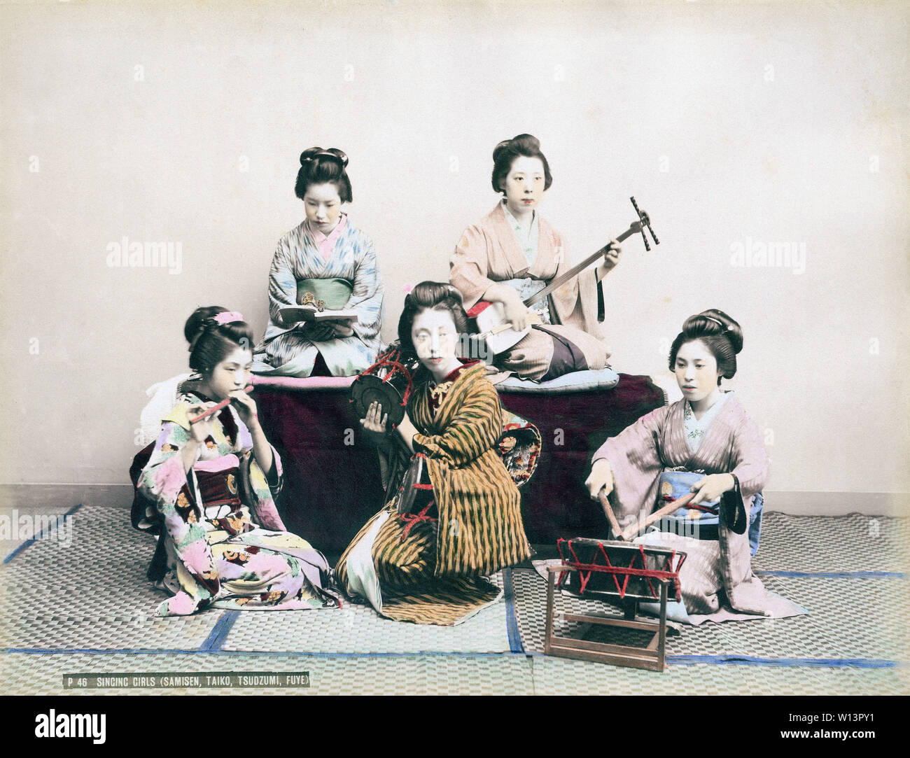 [ 1890s Japan - Maiko Playing Music ] —   Maiko in kimono and traditional hairstyles are practicing singing as well as playing tsutsumi (also tsuzumi) shoulder drums, shamisen, a three-stringed musical instrument, a taiko drum known as ootsutsumi and a fue flute.  19th century vintage albumen photograph. Stock Photo