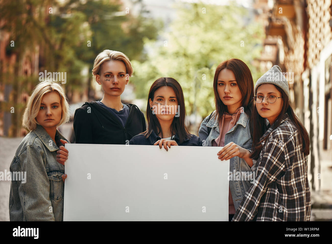 Group of protesting young female activists holding blank signboard and looking at camera while standing on the road. Women's march. Human rights. Protest concept Stock Photo