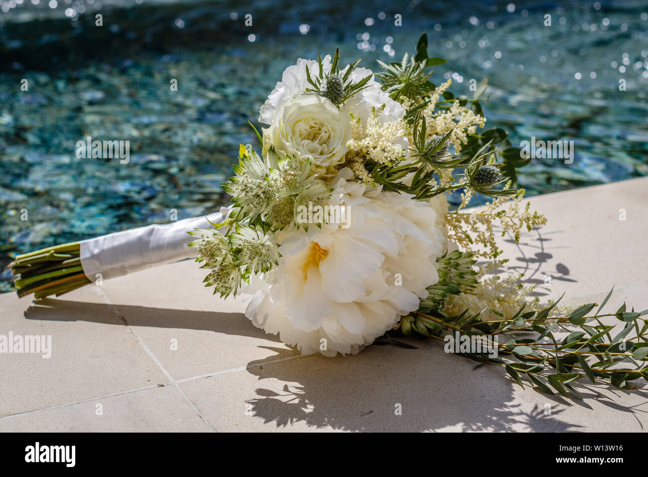Bali Roses Over Pool Wedding bouquet with white peony and roses laying on the edge of a ...
