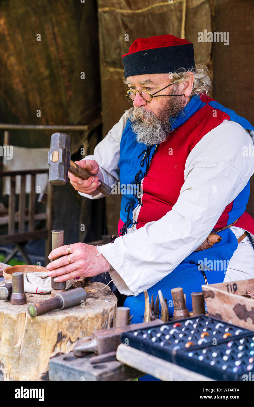 Medieval reenactment event. Senior man, metal worker, with bushy beard, sitting at work bench producing coins with hammer and wooden embossed stamp. Stock Photo