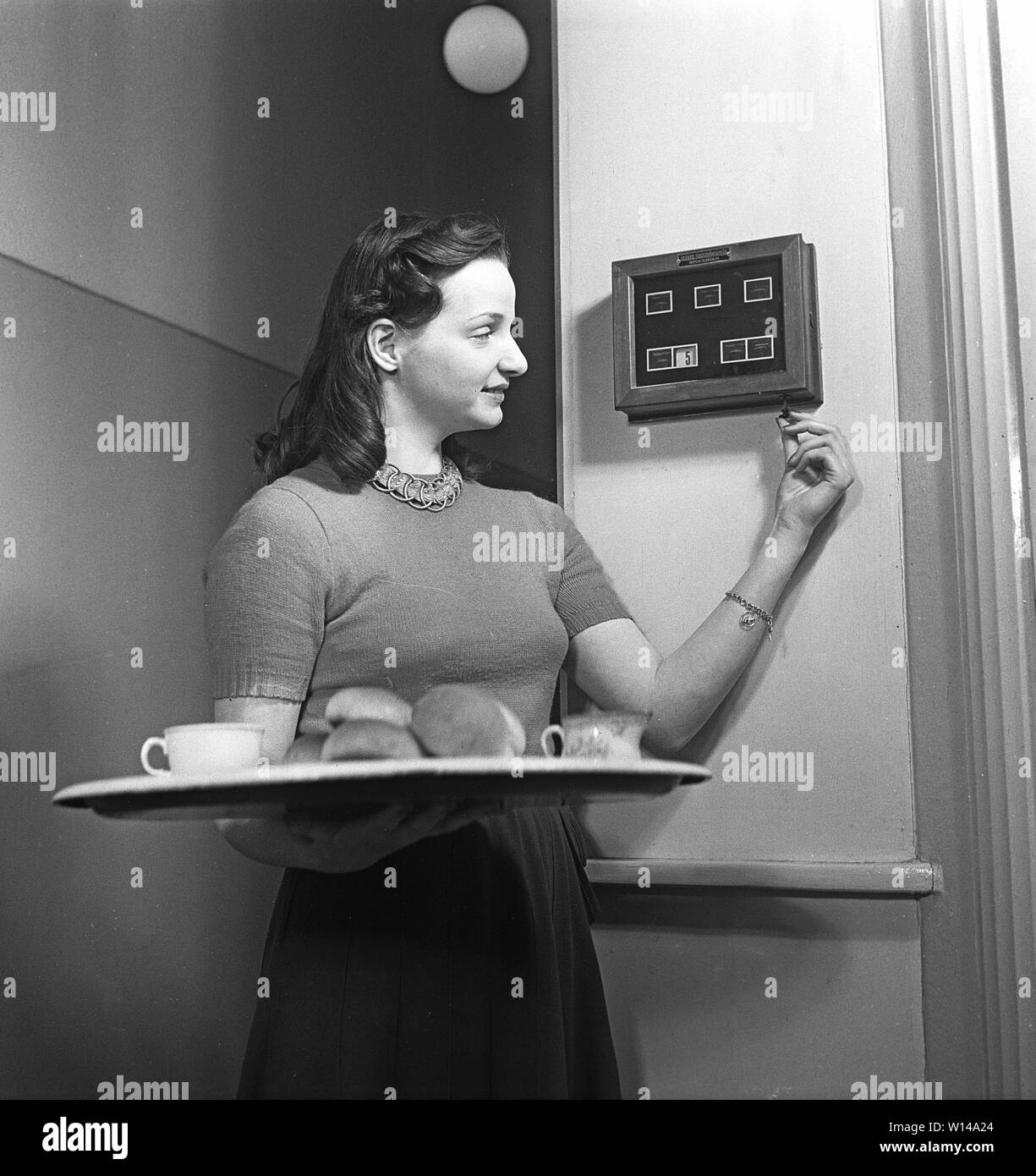 Service in the 1940s. A woman working as a maid or in a hotel is standing ready with a tray. A message board on the wall indicates where she goes with it. Number five could be the living room or another place in the building where someone has called upon attention. Sweden 1949 ref AN25-6 Stock Photo