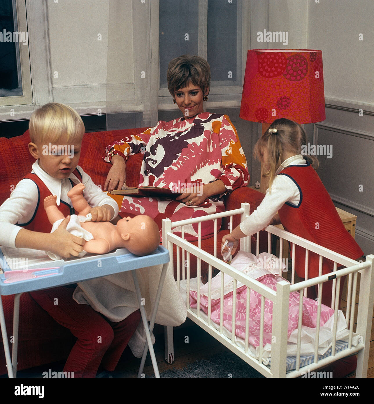 1960s mother. A young woman is reading a book while her children are playing together with dolls. The boy plays with a doll and puts clothes on it. The 60s was the decade of equality. Sweden 1964 Stock Photo