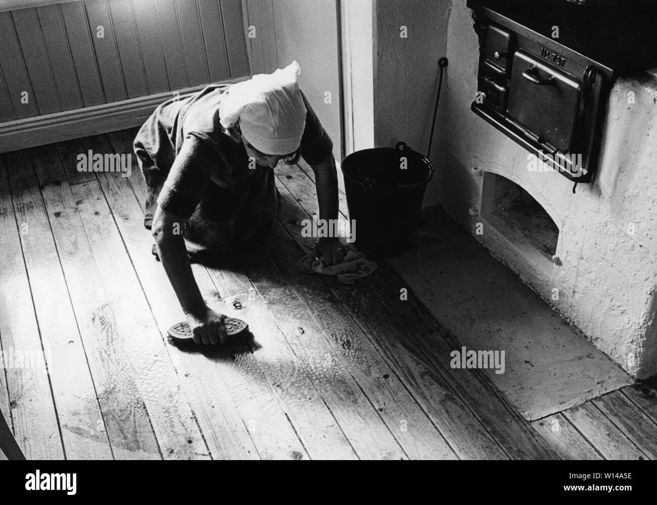 Cleaning the floor back then. An old woman is lying on the floor and use a scrubbing brush to get the kitchen floor clean. An old wooden kitchen stove is seen. Stock Photo
