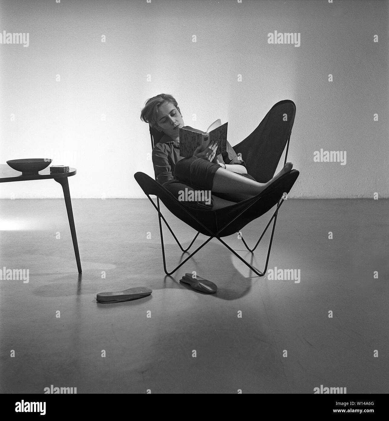 Home decor of the 1950s. A young woman is sitting in the  popular Butterfly chair, also known as a BKF chair or Hardoy chair. The chair has its nickname due to the design. For many people the chair is a symbol of the youth rebellion against a generation of parents that wanted their children to sit up straight. The chair was created by two argentinian architects Juan Kurchan and Jorge Ferrari Hardoy, and spanish Antonio Bonet in the 1930s. It was named BKD but was considered hard to sell, probably because of its futuristic design. American Hans Knoll re introduced it 1947 and it became an inter Stock Photo