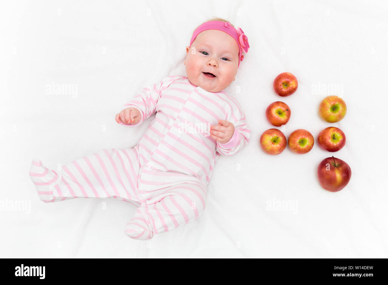 Cute little baby girl laying on white background with fresh red-green apples in shape of number four Stock Photo